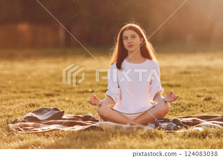 Doing yoga on the mat. Young girl is on the field at sunny daytime having nice weekend Doing yoga on the mat. Young girl is on the field at sunny daytime having nice weekend 124083038