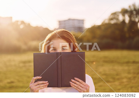 Holding book and looking into the camera. Young girl is on the field at sunny daytime having nice weekend Holding book and looking into the camera. Young girl is on the field at sunny daytime having nice weekend 124083092