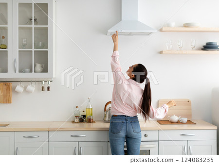 Household Concept. Back rear view of casual lady choosing and selecting mode on range exhaust hood, pushing button on mechanical fan above the stove. Female standing cooking in modern kitchen at home Household Concept. Back rear view of casual lady choosing and selecting mode on range exhaust hood, pushing button on mechanical fan above the stove. Female standing cooking in modern kitchen at home 124083235