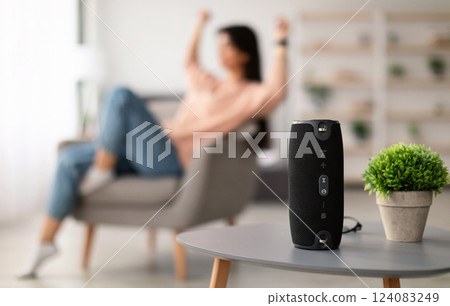 Modern Technology Concept. Closeup of smart portable wireless speaker on the table, selective focus. Young woman listening to music and dancing, sitting on armchair in the blurred background Modern Technology Concept. Closeup of smart portable wireless speaker on the table, selective focus. Young woman listening to music and dancing, sitting on armchair in the blurred background 124083249