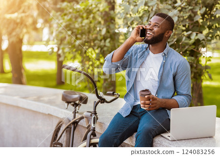 Freelance Lifestyle. Joyful Black Guy Talking On Cellphone And Drinking Takeaway Coffee While Working On Laptop Outdoors, His Bike Parked Next By 124083256