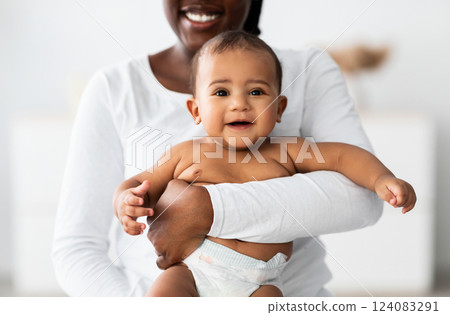 Mother's Happiness. Closeup portrait of positive black baby, young African American woman hugging and holding on hands her pretty small kid in diaper standing in bedroom at home. Childcare Concept 124083291
