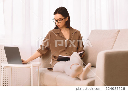 Checking Work Schedule. Focused young woman in specs lying on the couch with notebook, using laptop, copyspace Checking Work Schedule. Focused young woman in specs lying on the couch with notebook, using laptop, copyspace 124083326