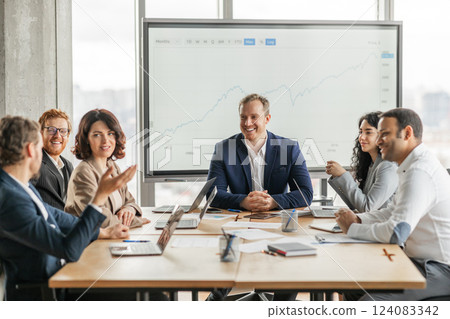 A group of business professionals sit around a large wooden conference table, engaged in a meeting. They are looking at a large screen behind them displaying a projected growth graph A group of business professionals sit around a large wooden conference table, engaged in a meeting. They are looking at a large screen behind them displaying a projected growth graph 124083342