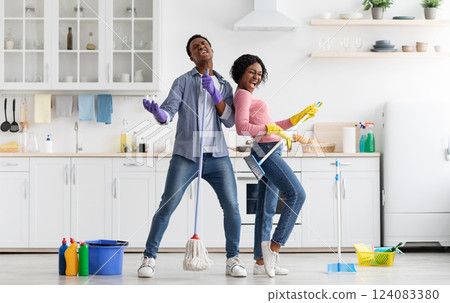 Joyful black loving couple singing songs while cleaning kitchen, using broom and mop as microphones and guitar, cheerful african american young man and woman imitating rock stars while house-keeping Joyful black loving couple singing songs while cleaning kitchen, using broom and mop as microphones and guitar, cheerful african american young man and woman imitating rock stars while house-keeping 124083380
