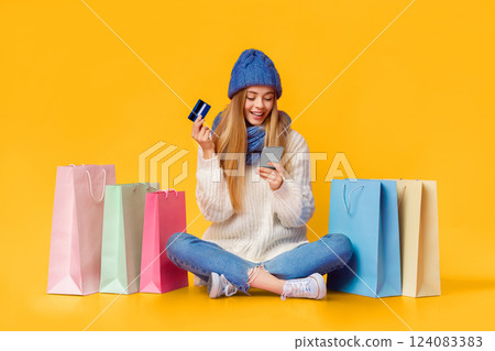 Pretty blonde girl in blue knitted hat using smartphone and credit card for online shopping, sitting on floor among shopping bags, pink background, copy space Pretty blonde girl in blue knitted hat using smartphone and credit card for online shopping, sitting on floor among shopping bags, pink background, copy space 124083383