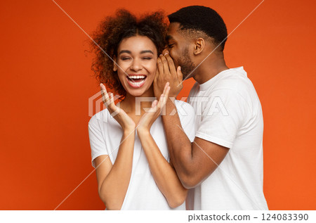 African-american girl laughing, listening how her boyfriend whispering secret or gossips, orange studio background African-american girl laughing, listening how her boyfriend whispering secret or gossips, orange studio background 124083390