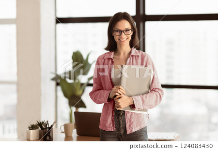 Happy Worker. Smiling woman in eyeglasses holding document case, standing next to desk, looking at camera, copyspace 124083430