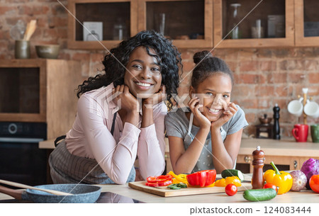 African sweet mom and daughter leaning on elbows and smiling, cooking healthy meal together at kitchen, free space African sweet mom and daughter leaning on elbows and smiling, cooking healthy meal together at kitchen, free space 124083444