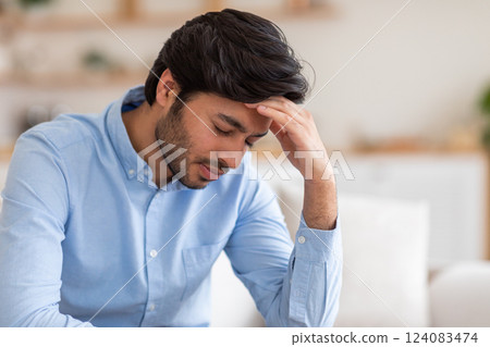 Arab man with dark hair is sitting in a chair indoors. He has a stressed expression on his face. He is holding his hand to his forehead, as if he is experiencing a headache or feeling overwhelmed. 124083474