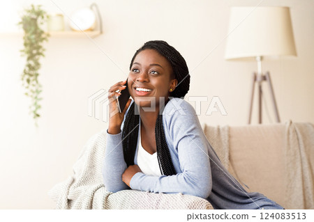 Young african woman talking on mobile phone with friend while sitting on comfortable couch at home, copy space 124083513