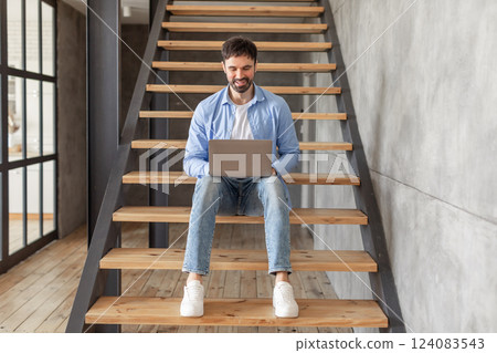 A man wearing a blue shirt and jeans sits on the wooden stairs in a modern home. He is working on a laptop computer, smiling. The stairway has a metal railing and leads to an upper level of the home. A man wearing a blue shirt and jeans sits on the wooden stairs in a modern home. He is working on a laptop computer, smiling. The stairway has a metal railing and leads to an upper level of the home. 124083543