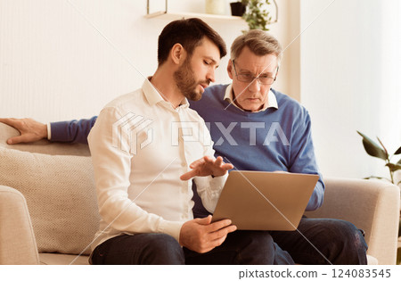 Digital Literacy. Middle-Aged Son Teaching Elderly Father To Use Laptop Computer, Social Media And Internet Sitting On Couch Indoor 124083545