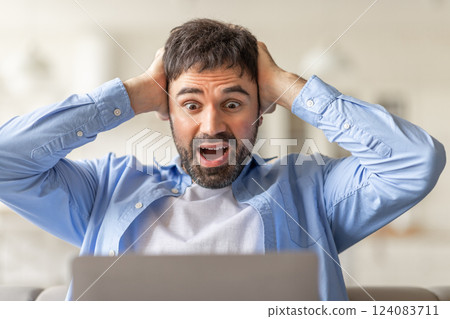 A man sits in front of a laptop, staring at the screen with shock and surprise. He holds his head in his hands with a wide-eyed expression and an open mouth, closeup 124083711