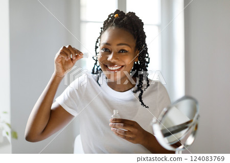 A young black woman with braided hair is applying facial serum to her face. She is smiling and looking at the mirror in front of her. She is wearing a white t-shirt and is sitting in front of window. 124083769