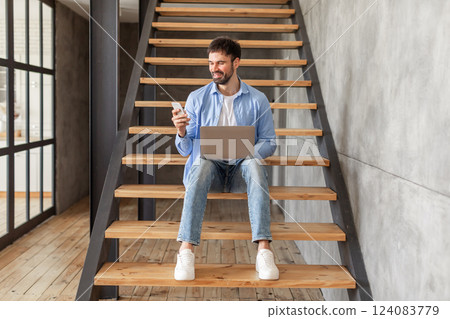 A man sits on a set of wooden stairs in a modern home, using his phone and a laptop. He is smiling, looking down at his phone, while holding the laptop on his lap. 124083779
