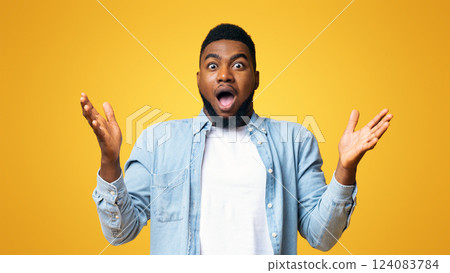 Portrait of surprised african american man standing with opened mouth and raised hands, yellow studio background 124083784