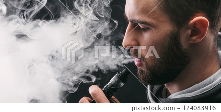 Man with vaping mod exhaling steam at black studio background. Bearded guy smoking e-cigarette to quit tobacco. Vapor and alternative nicotine free smoking concept, closeup Man with vaping mod exhaling steam at black studio background. Bearded guy smoking e-cigarette to quit tobacco. Vapor and alternative nicotine free smoking concept, closeup 124083916