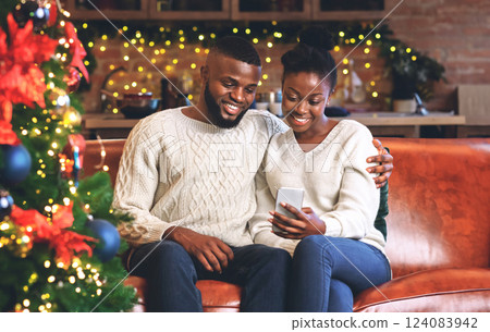 Portrait of smiling african couple using smartphone, reading Christmas wishes from friends while sitting on couch near Xmas tree at home. 124083942