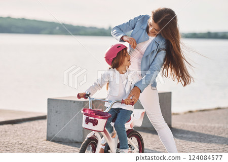 Woman is helping the little girl. Mother with her young daughter is with bicycle outdoors together Woman is helping the little girl. Mother with her young daughter is with bicycle outdoors together 124084577