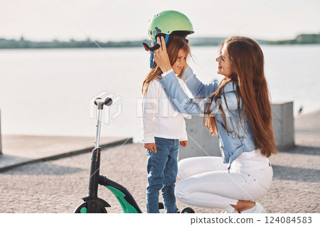 In green protective helmet. Cute little girl with scooter is outdoors in summertime 124084583
