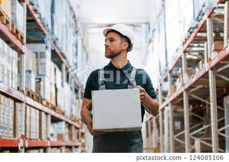 Walking with white box in hands. Employee in uniform is working in the storage at daytime Walking with white box in hands. Employee in uniform is working in the storage at daytime 124085106
