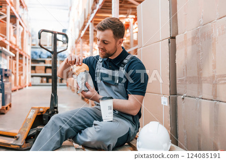 Man is eating lunch. Taking a break. Employee in uniform is working in the storage at daytime Man is eating lunch. Taking a break. Employee in uniform is working in the storage at daytime 124085191