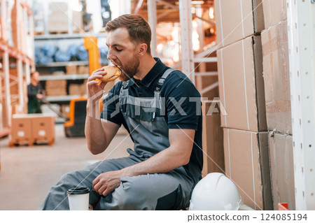 Man is eating lunch. Taking a break. Employee in uniform is working in the storage at daytime Man is eating lunch. Taking a break. Employee in uniform is working in the storage at daytime 124085194