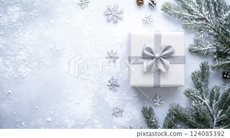 A flat lay photograph featuring a white gift box with a silver bow, surrounded by evergreen branches with a light dusting of snow 124085392
