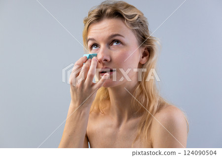 A Thoughtful Young Woman Applying Cosmetic Products to Her Face for an Everyday Makeup Routine in a Light Grey Background 124090504