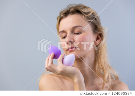A Woman Contemplates Beauty Tools: Exploring Makeup Sponge Usage with a Thoughtful Look and Soft Expression Under Natural Light Against a Light Gray Background A Woman Contemplates Beauty Tools: Exploring Makeup Sponge Usage with a Thoughtful Look and Soft Expression Under Natural Light Against a Light Gray Background 124090554