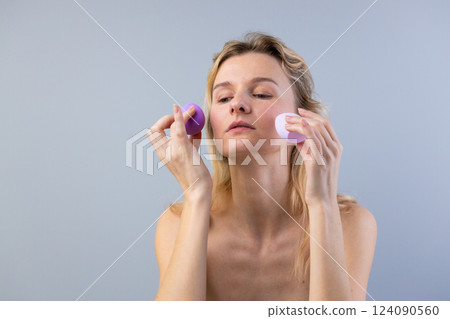 Aesthetic Beauty Scene Featuring a Young Woman Applying Makeup with Purple Sponges Against a Soft Gray Background for a Serene Atmosphere Aesthetic Beauty Scene Featuring a Young Woman Applying Makeup with Purple Sponges Against a Soft Gray Background for a Serene Atmosphere 124090560