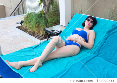 Relaxing by the Pool: A Sun-Kissed Woman in a Bikini and Sunglasses Enjoys the Warm Summer Day on a Blue Towel, Embracing the Joy of Leisure and Tranquility Under the Clear Sky, Surrounded by the Relaxing by the Pool: A Sun-Kissed Woman in a Bikini and Sunglasses Enjoys the Warm Summer Day on a Blue Towel, Embracing the Joy of Leisure and Tranquility Under the Clear Sky, Surrounded by the 124090833
