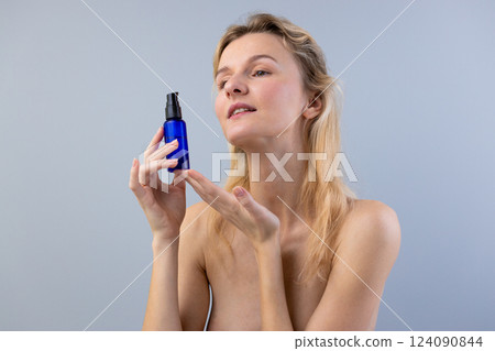 Exploring the Beauty of Skincare: A Woman Holding a Blue Cosmetic Bottle with Radiance in Her Eyes Against a Soft Gray Background Exploring the Beauty of Skincare: A Woman Holding a Blue Cosmetic Bottle with Radiance in Her Eyes Against a Soft Gray Background 124090844