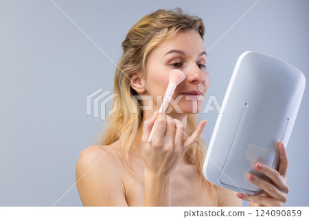 A Young Woman Applying Makeup with a Soft Brush While Looking in a Mirror: Embracing Beauty and Self-Care in a Calm and Simple Environment 124090859