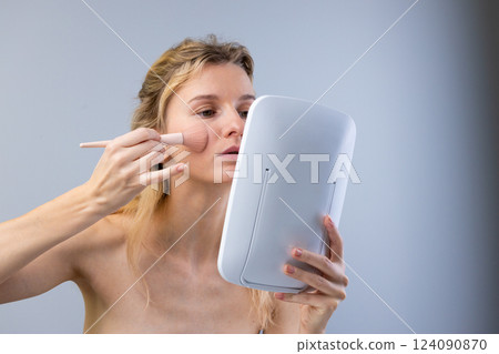A woman applying makeup with a brush in front of a mirror, focusing on her cheek, highlighting the beauty routine and self-care approach in a serene atmosphere. 124090870