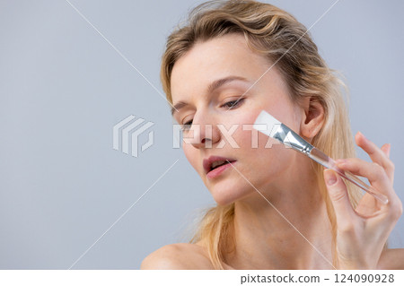 Woman with blonde hair applying skincare products using a brush, showcasing a beauty routine focused on facial care and enhancing natural features in a soft light 124090928