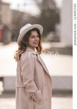 A woman wearing a white hat and a tan coat is standing in front of a building. She has a smile on her face and she is enjoying the moment. A woman wearing a white hat and a tan coat is standing in front of a building. She has a smile on her face and she is enjoying the moment. 124091583