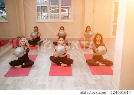 A group of six athletic women doing pilates or yoga on pink mats in front of a window in a beige loft studio interior. Teamwork, good mood and healthy lifestyle concept. 124091614