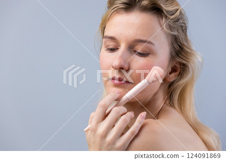 A Young Woman Applying Makeup with a Soft Blush Brush on Her Cheek Against a Soft Gray Background A Young Woman Applying Makeup with a Soft Blush Brush on Her Cheek Against a Soft Gray Background 124091869
