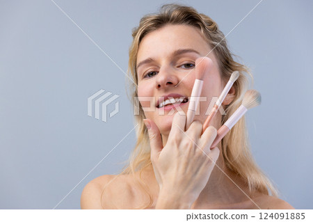 Portrait of a Smiling Woman Holding Beauty Brushes Against a Soft Gray Background, Showcasing the Art of Makeup Application and the Joy of Self-Care 124091885