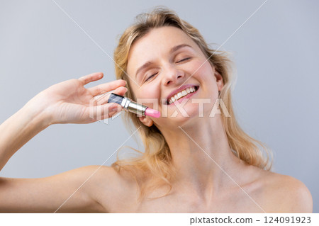 Joyful Woman Enjoying a Pink Lipstick while Smiling and Showing Happiness in a Studio Setting with a Neutral Background 124091923