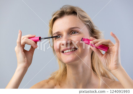 A young woman cheerfully applying mascara in a bright studio, showcasing a joyful moment in her beauty routine while highlighting her striking features and genuine smile. A young woman cheerfully applying mascara in a bright studio, showcasing a joyful moment in her beauty routine while highlighting her striking features and genuine smile. 124091993