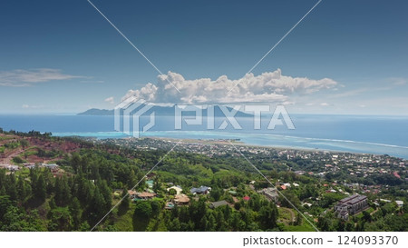 Aerial view of Moorea island in horizon with its characteristic mountains partially covered by clouds under blue sky, seen from Tahiti, with its airport and residential areas with lush vegetation. 124093370