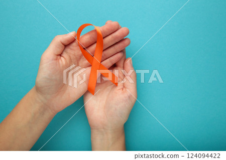 Hands holding orange color ribbon on blue background. Kidney Cancer Awareness, Leukemia disease, Skin cancer awareness, World Cancer Day. Hands holding orange color ribbon on blue background. Kidney Cancer Awareness, Leukemia disease, Skin cancer awareness, World Cancer Day. 124094422