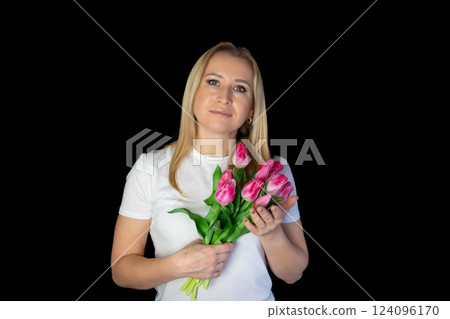 Happy blonde woman in white t-shirt holding bunch of tulips while standing against black background 124096170