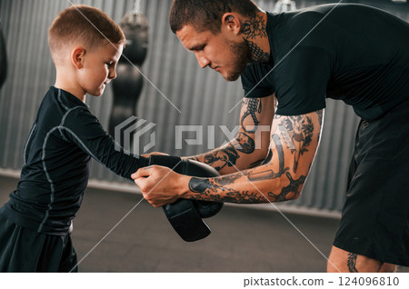 Man is helping child to wear the glowes. Coach is teaching the boy box techniques indoors Man is helping child to wear the glowes. Coach is teaching the boy box techniques indoors 124096810