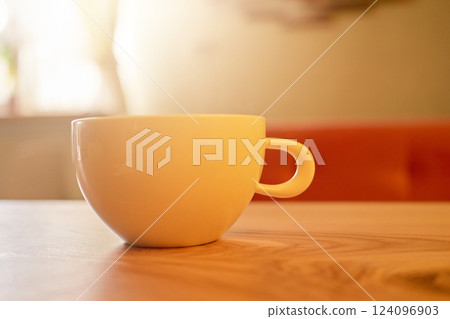 A ceramic cup rests on a smooth wooden table, illuminated by soft afternoon light. The warm ambiance suggests a perfect spot for enjoying beverages and relaxation 124096903
