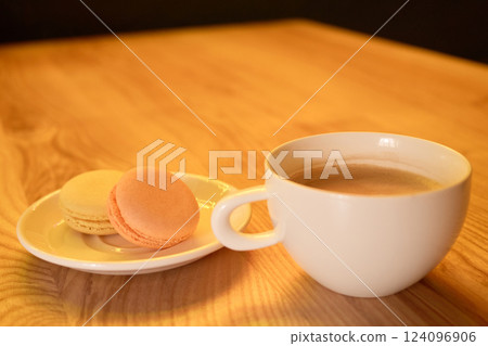 A warm beverage sits in a white cup next to a plate with two colorful macarons on a smooth wooden table, capturing a cozy afternoon moment at a cafe A warm beverage sits in a white cup next to a plate with two colorful macarons on a smooth wooden table, capturing a cozy afternoon moment at a cafe 124096906