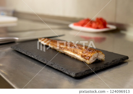 A perfectly grilled fish filet rests on a sleek black slate plate. In the background, vibrant red ingredients are prepared, showcasing a lively kitchen atmosphere A perfectly grilled fish filet rests on a sleek black slate plate. In the background, vibrant red ingredients are prepared, showcasing a lively kitchen atmosphere 124096913